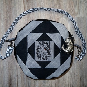 Könnte beinhalten: Eine schwarze und graue runde Tasche mit einem Wolf-Design und einem silbernen Drachen-Anhänger. Die Tasche hat einen schwarz-weißen gemusterten Riemen.