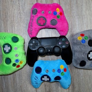 Könnte beinhalten: Vier Plüsch-Spielcontroller in Grün, Pink, Blau und Grau, mit den Namen "Janik", "Alexandra", "Julia" und "Geert" bestickt. Ein schwarzer PlayStation 4-Controller befindet sich in der Mitte.