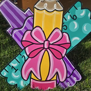 May include: A wooden craft project featuring a purple crayon, a yellow pencil with a pink bow, and a teal ruler with white polka dots and numbers 1, 2, 5, and 6.