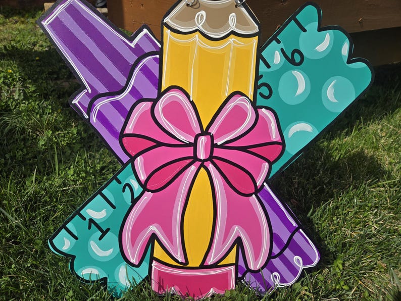 May include: A wooden craft project featuring a purple crayon, a yellow pencil with a pink bow, and a teal ruler with white polka dots and numbers 1, 2, 5, and 6.