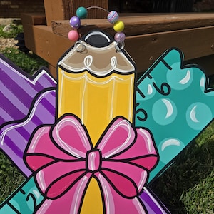 May include: A wooden door hanger decorated with a pencil, ruler, and marker. The pencil is yellow with a brown eraser and a pink bow. The ruler is teal with white circles. The marker is purple with white stripes. The hanger is decorated with a colorful beaded garland.