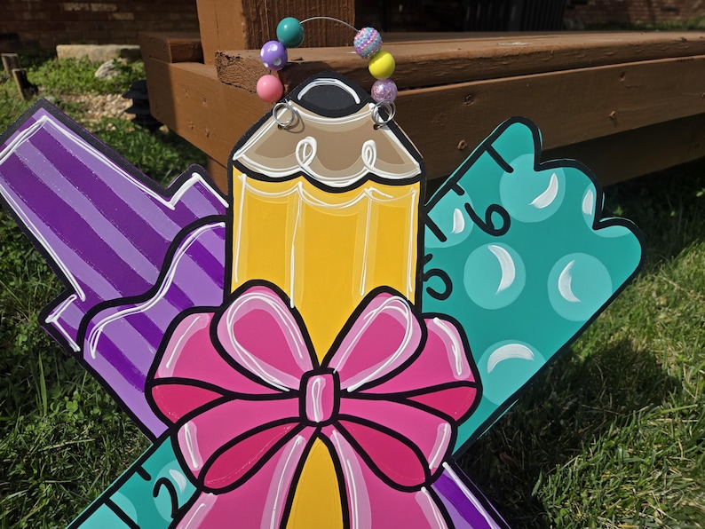 May include: A wooden door hanger decorated with a pencil, ruler, and marker. The pencil is yellow with a brown eraser and a pink bow. The ruler is teal with white circles. The marker is purple with white stripes. The hanger is decorated with a colorful beaded garland.