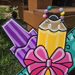 May include: A wooden back-to-school sign with a purple marker, a yellow pencil with a pink bow, and a green ruler with white numbers. The sign is decorated with colorful beads.