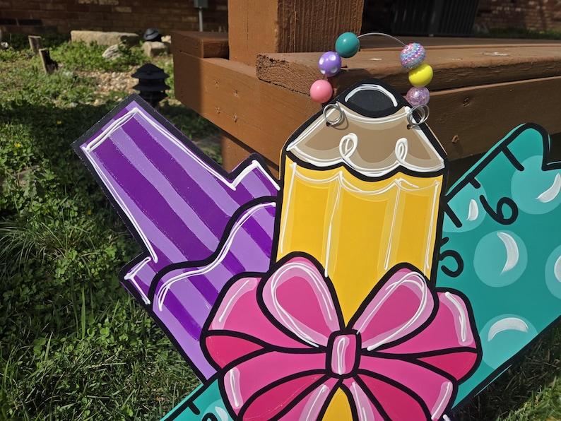 May include: A wooden back-to-school sign with a purple marker, a yellow pencil with a pink bow, and a green ruler with white numbers. The sign is decorated with colorful beads.