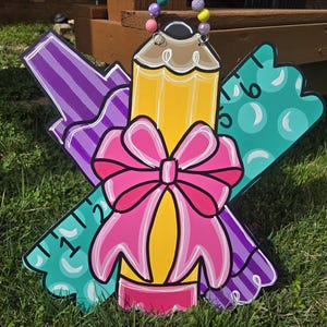 May include: A wooden door hanger shaped like a pencil, ruler, and marker with a pink bow. The pencil is yellow, the ruler is teal, and the marker is purple.
