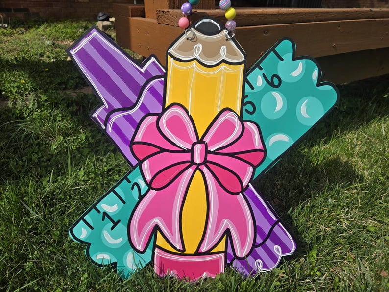May include: A wooden door hanger shaped like a pencil, ruler, and marker with a pink bow. The pencil is yellow, the ruler is teal, and the marker is purple.