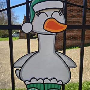 May include: A whimsical, cartoon-style goose decoration. The goose is primarily white and gray, with an orange beak and green hat and shoes. The hat has a white brim and a white pom-pom. The decoration is hanging on a black metal fence.
