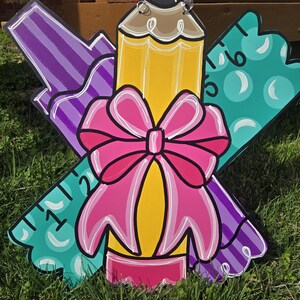 May include: A wooden door hanger decorated with a pencil, a ruler, and a marker. The pencil is yellow with a pink eraser and a brown tip. The ruler is green with white numbers. The marker is purple with white stripes. A pink bow is tied around the pencil.
