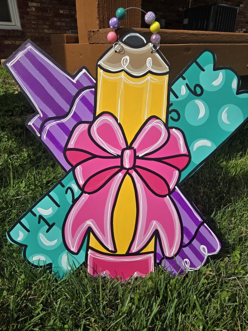 May include: A wooden door hanger decorated with a pencil, a ruler, and a marker. The pencil is yellow with a pink eraser and a brown tip. The ruler is green with white numbers. The marker is purple with white stripes. A pink bow is tied around the pencil.