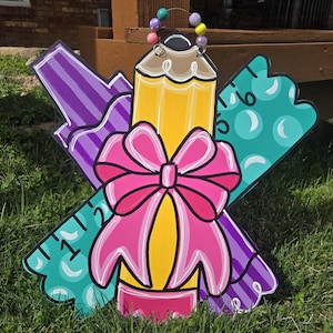 May include: A wooden door hanger decorated with a pencil, a ruler, and a crayon, all in bright colors. A pink bow is tied around the pencil. The door hanger is perfect for a classroom or a child's room.