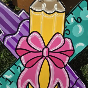 May include: A wooden craft project featuring a yellow pencil, a purple marker, and a teal ruler. The pencil has a pink bow tied around it. The craft is decorated with white outlines and has a back-to-school theme.