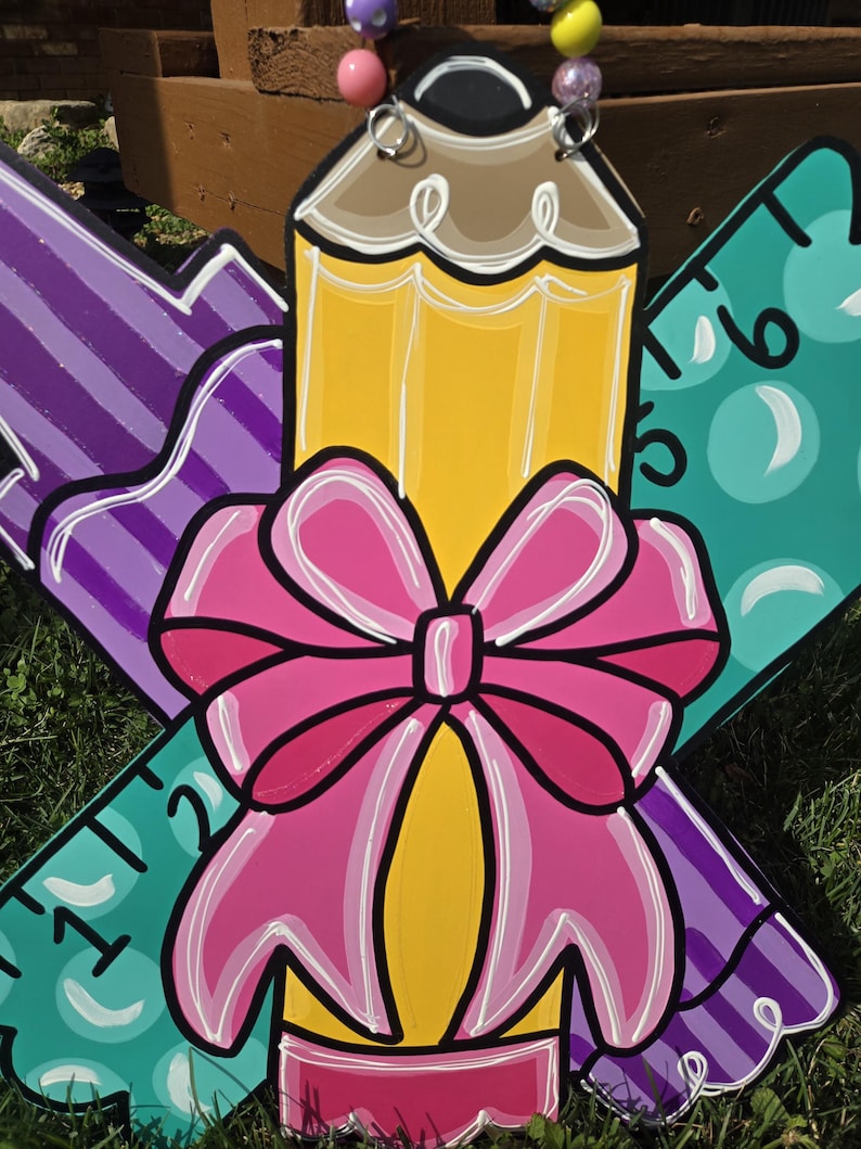 May include: A wooden craft project featuring a yellow pencil, a purple marker, and a teal ruler. The pencil has a pink bow tied around it. The craft is decorated with white outlines and has a back-to-school theme.