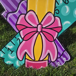 May include: A wooden cutout of a pencil with a pink bow, a purple ruler, and a green ruler with a bubble pattern. The pencil is yellow with a pink eraser. The rulers have numbers on them.