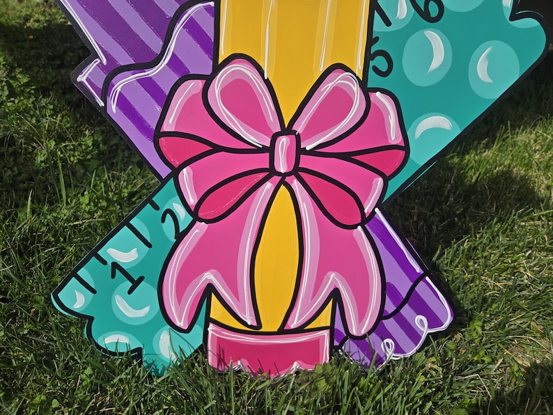 May include: A wooden cutout of a pencil with a pink bow, a purple ruler, and a green ruler with a bubble pattern. The pencil is yellow with a pink eraser. The rulers have numbers on them.