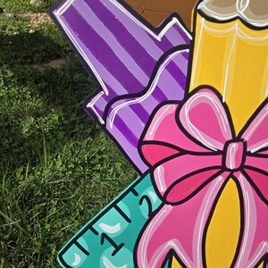 May include: A colorful wooden cutout of school supplies, including a purple marker, a yellow pencil with a pink bow, and a turquoise ruler with the numbers "1" and "2" visible.
