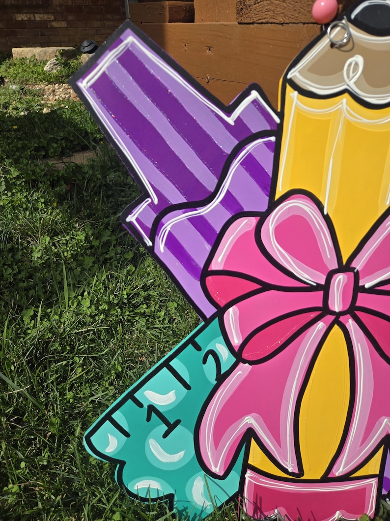 May include: A colorful wooden cutout of school supplies, including a purple marker, a yellow pencil with a pink bow, and a turquoise ruler with the numbers "1" and "2" visible.