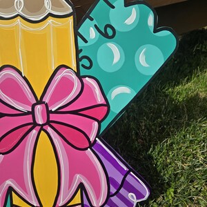 May include: A wooden cutout of a yellow pencil with a pink bow, a purple and white striped ribbon, and a teal ruler with white polka dots. The cutout is painted in a cartoon style.