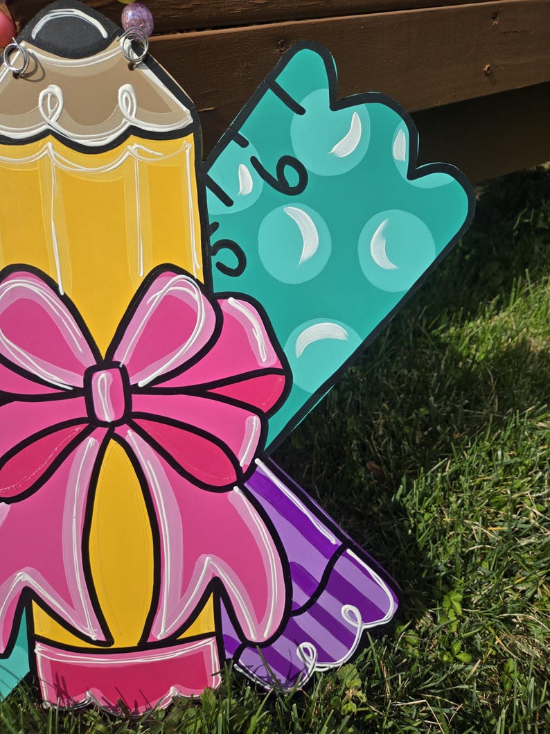 May include: A wooden cutout of a yellow pencil with a pink bow, a purple and white striped ribbon, and a teal ruler with white polka dots. The cutout is painted in a cartoon style.