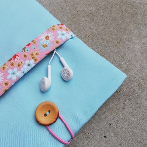 Może przedstawiać: Jasno błękitna torba z tkaniny z różową ozdobą z kwiatowym nadrukiem. Torba ma drewniany guzik i różową gumkę. Białe słuchawki wystają z torby.
