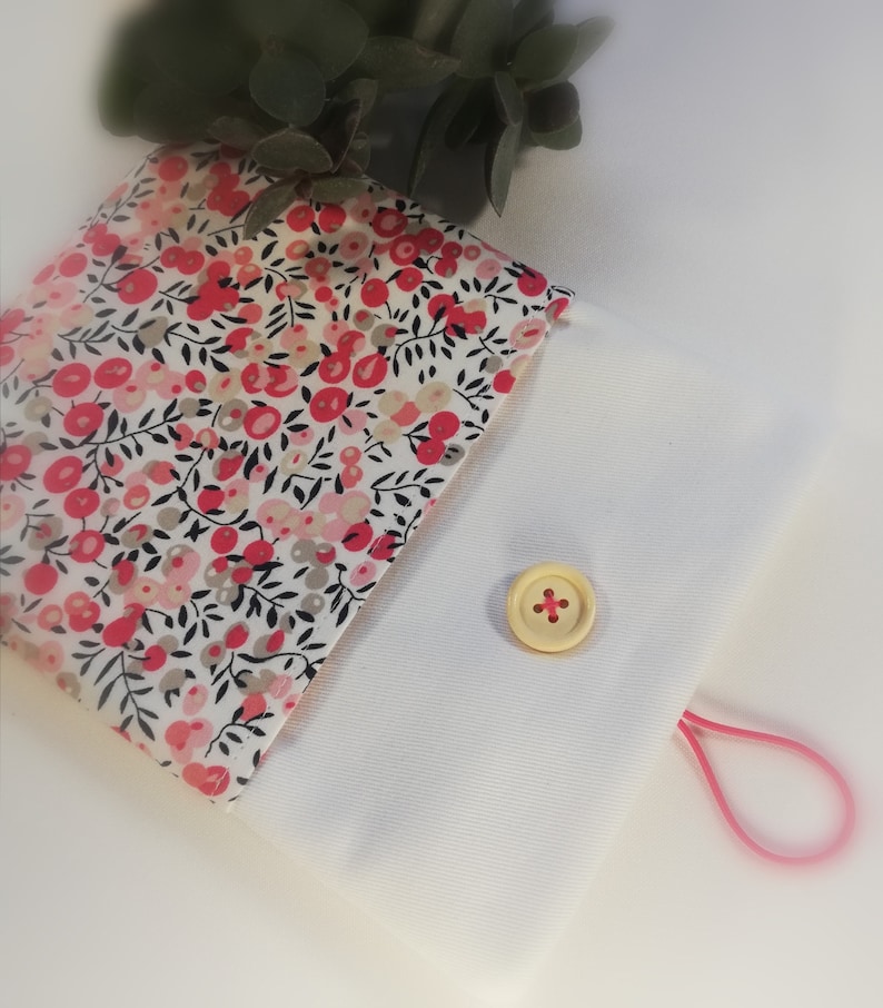 Puede incluir: Una peque&ntilde;a bolsa blanca con un estampado floral rosa en la solapa delantera. La bolsa tiene un cierre de bot&oacute;n &uacute;nico y un lazo el&aacute;stico rosa.