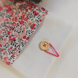 Puede incluir: Una bolsa de tela blanca con un forro de estampado floral. La bolsa tiene una banda el&aacute;stica rosa y un cierre de bot&oacute;n de madera.