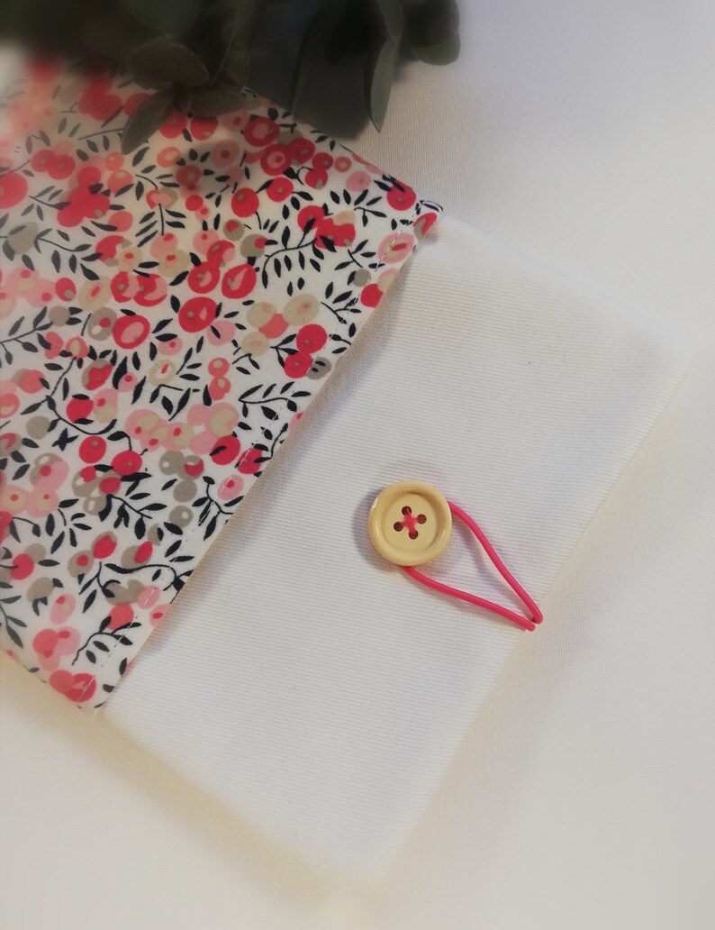 Puede incluir: Una bolsa de tela blanca con un forro de estampado floral. La bolsa tiene una banda el&aacute;stica rosa y un cierre de bot&oacute;n de madera.