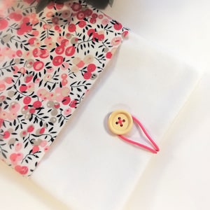 Puede incluir: Una bolsa de tela blanca con estampado floral rosa y cierre de bot&oacute;n de madera. La bolsa se cierra con una banda el&aacute;stica rosa.