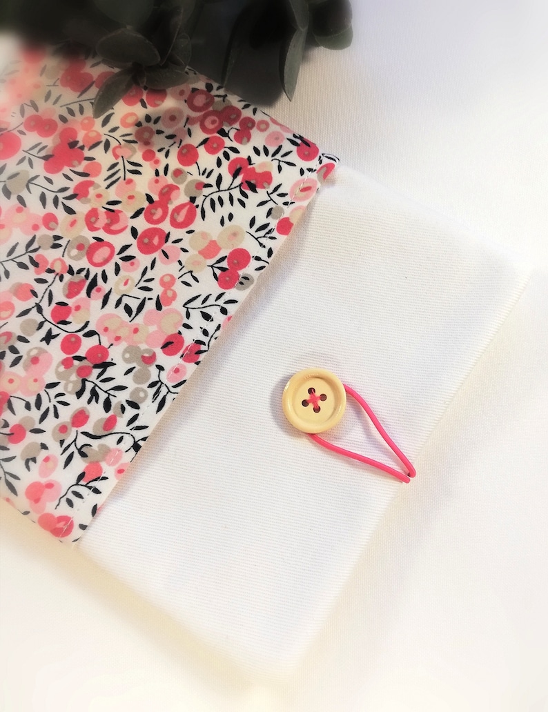 Puede incluir: Una bolsa de tela blanca con estampado floral rosa y cierre de bot&oacute;n de madera. La bolsa se cierra con una banda el&aacute;stica rosa.