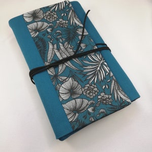 Peut inclure: Un carnet en tissu bleu sarcelle avec un imprimé floral noir et blanc. Le carnet est fermé par un cordon noir.