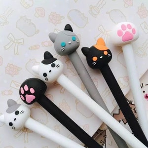 K&ouml;nnte beinhalten: Sechs Stifte mit verschiedenen Katzenkopf-Toppern. Die Topper sind in verschiedenen Farben, darunter Schwarz, Wei&szlig;, Grau, Orange und Pink. Die Stifte sind alle wei&szlig; mit schwarzer Tinte.