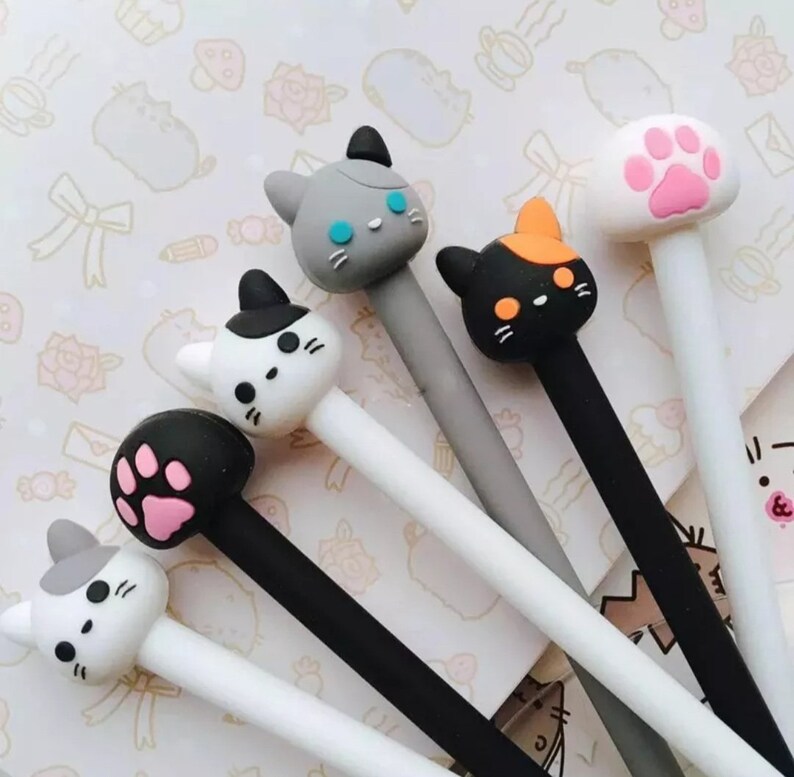 K&ouml;nnte beinhalten: Sechs Stifte mit verschiedenen Katzenkopf-Toppern. Die Topper sind in verschiedenen Farben, darunter Schwarz, Wei&szlig;, Grau, Orange und Pink. Die Stifte sind alle wei&szlig; mit schwarzer Tinte.