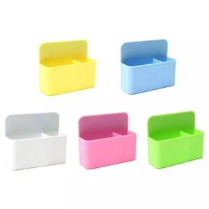 May include: A collection of five colorful plastic organizers. Each organizer has a rectangular body with a divided interior and a hinged lid. The colors include yellow, blue, white, pink, and green. The organizers are designed for desk or storage use.