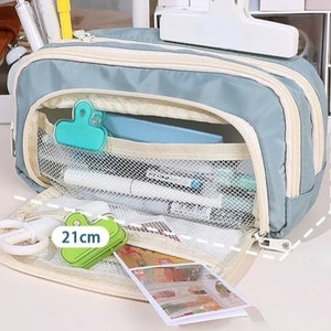 May include: A dusty blue pencil case with cream-colored zippers and trim. The interior features a white mesh pocket containing pens and a teal binder clip. A green binder clip is visible near a label that reads 21cm, indicating the length.