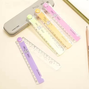 May include: Four folding rulers in pastel purple, yellow, pink, and light green, each featuring a small flower-shaped knob at the hinge and the text "STUDY TIME!" with a cartoon cat illustration. A grey metal pencil case and a wooden pencil are visible.