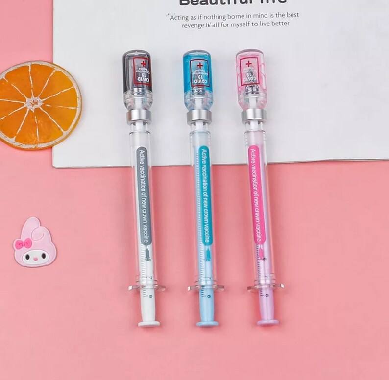 May include: Three syringe-shaped pens with clear barrels and colorful tops. The pens have black, blue, and pink accents, with text that reads "COVID 19" and "Active vaccination of new crown vaccine". The pens are on a pink surface.