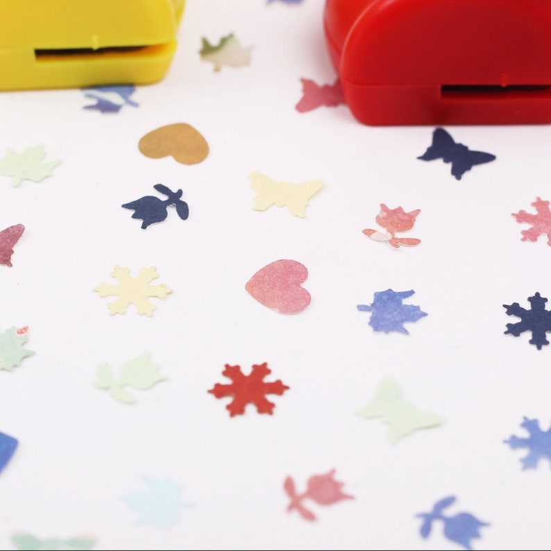 May include: Two colorful paper punch tools with different designs. The tools are on a white surface with confetti cutouts in various shapes and colors, including hearts, butterflies, snowflakes, and leaves.