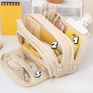 May include: A cream-colored multi-compartment pencil case with three zippered sections. The case is open, revealing organized stationery including pens, yellow notebooks, and sticky notes. A large white clip is attached to the top right side.
