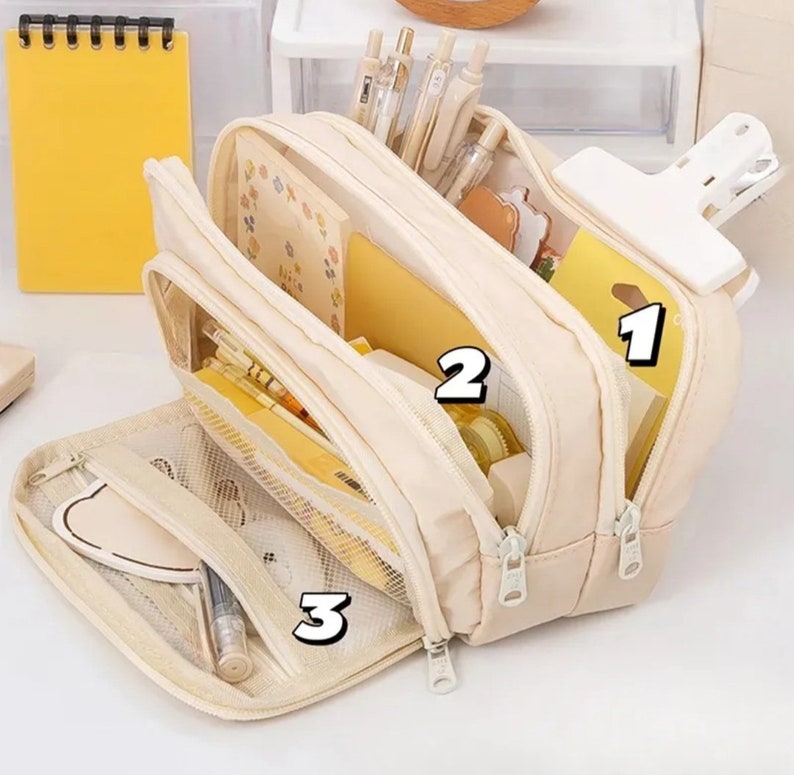 May include: A cream-colored multi-compartment pencil case with three zippered sections. The case is open, revealing organized stationery including pens, yellow notebooks, and sticky notes. A large white clip is attached to the top right side.
