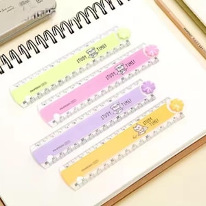May include: Four plastic rulers in light green, pink, purple, and yellow, each featuring a small flower-shaped topper, a cat illustration, and the text "STUDY TIME!" printed on the surface. The rulers display clear black measurement markings.