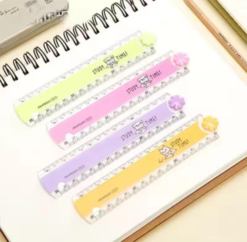 May include: Four plastic rulers in light green, pink, purple, and yellow, each featuring a small flower-shaped topper, a cat illustration, and the text "STUDY TIME!" printed on the surface. The rulers display clear black measurement markings.