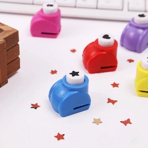 May include: Five colorful paper punchers with different shapes: a star, a heart, a flower, a circle, and a square. The punchers are on a white surface with confetti.
