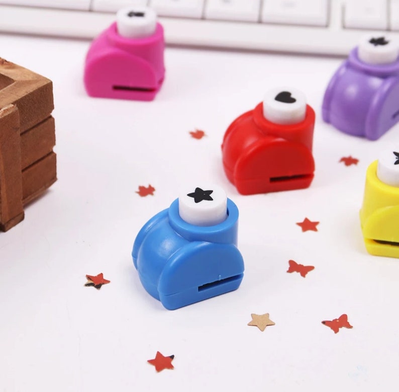 May include: Five colorful paper punchers with different shapes: a star, a heart, a flower, a circle, and a square. The punchers are on a white surface with confetti.
