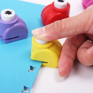 May include: A close-up of a yellow butterfly shaped paper punch being used to punch a blue piece of paper. The punch has a white button on top and is being pressed down with a finger. There are two other punches in the background, one purple and one red.