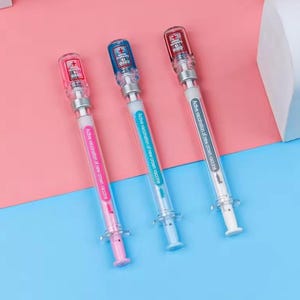 May include: Three syringe-shaped pens with clear barrels and colorful accents. Each pen has a different colored plunger: pink, blue, and red. The pens have the text "COVID 19" and "Active vaccination of new crown vaccine."