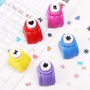 May include: Five colorful paper punchers with different designs. The designs include a flower, a heart, a butterfly, a leaf, and a snowflake. The punchers are on a white background with confetti.