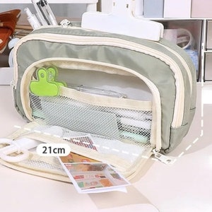 May include: A sage green pencil case with a cream-colored zipper and trim. The interior features a white mesh pocket, a lime green binder clip, and a white plastic clip on top. A label clearly displays the measurement of 21cm for the length.