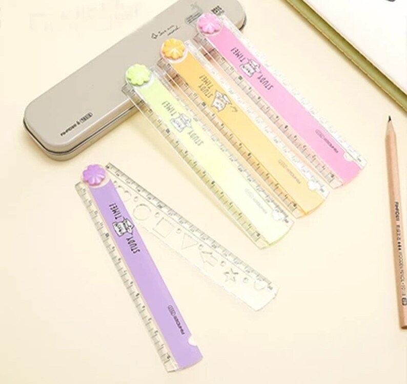 May include: A set of four plastic rulers in pastel purple, yellow, orange, and pink, each featuring a flower-shaped pivot, a cat illustration, and the text STUDY TIME. The rulers include stencil cutouts for shapes and metric measurement markings.