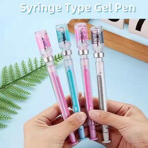 May include: Four gel pens designed to resemble syringes, with clear barrels and colorful tops. Each pen has a different colored ink and a small vial-shaped top with the word "COVID" printed on it. The text "Syringe Type Gel Pen" is at the top.