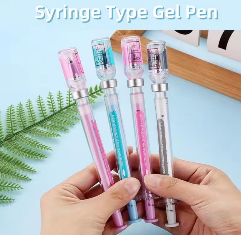 May include: Four gel pens designed to resemble syringes, with clear barrels and colorful tops. Each pen has a different colored ink and a small vial-shaped top with the word "COVID" printed on it. The text "Syringe Type Gel Pen" is at the top.