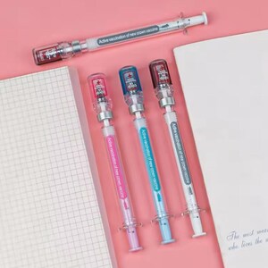 May include: A collection of syringe-shaped pens with colorful accents. The pens have clear barrels with colored plungers and caps, and the text "Active vaccination of new crown vaccine" is printed on the barrels. The pens are arranged on a pink surface.